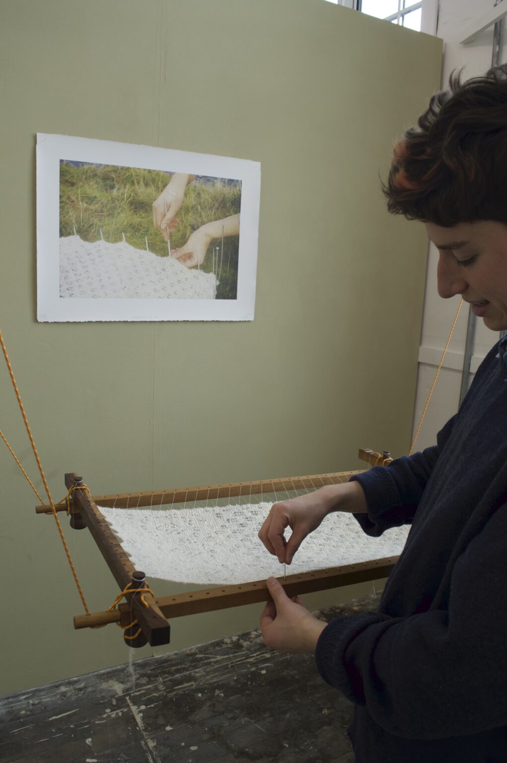 This hap frame hangs on thin, orange rope from a hand-made, hand-lathed, oak pulley system. It is like a pulley you might find on boat rigging, or a Glaswegian, tenemental washing airer. A piece of knitted lace is stretched in the middle of the hap frame. Behind the frame is hung a lithography print of a 35mm film photograph. The white paper is pinned at the four corners with sewing pins into cork behind. The image itself is set amongst the mossy, loch edge in the Kilpatrick hills, just outside Glasgow. The blue ripples of loch Humphrey are just visible on the top edge of the image. Two young, white hands touch the knitting and steel knitting needles – pinning it out on the grass and moss. Small moss sporophytes peek through some holes in the lace. There is a gentle light settled across the photo and small shadows of the knitting needles are cast on the hand closest to the foreground.