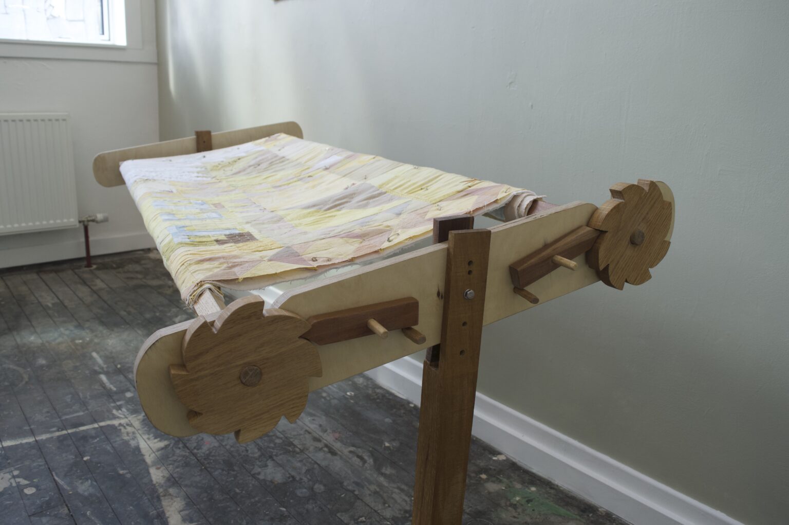 A quilt frame made of oak, cherry, and iroko wood stands in the space. The sturdy and delicate wooden structure has been sanded, oiled, and waxed so it is smooth and sumptuous to the touch. It has two legs with decoratively detailed feet. On one end it has two wooden cogs which help turn the beams and the quilt. A quilting frame is somewhat like a hollow table, with moving parts, to allow a quilt to be stretched across its limbs. Upon this frame a naturally dyed quilt is stretched.